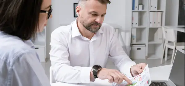Mężczyzna z zarostem i zegarkiem na ręku analizuje wykresy, podczas gdy kobieta w okularach obserwuje. Ich praca wymaga ścisłego nadzoru.