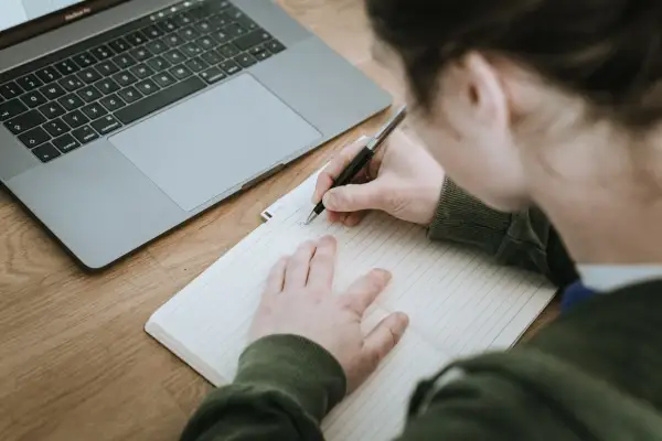 Nauczyciel pisze opinię o uczniu w zeszycie, obok leży laptop.