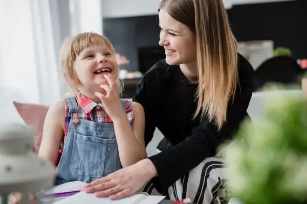 Uśmiechnięta dziewczynka z niepełnosprawnością sprzężoną bawi się z mamą, która wspiera ją w nauce.