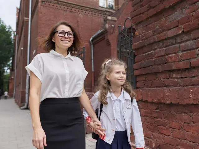 Uśmiechnięta mama z córką idą na rozpoczęcie roku szkolnego. Dziewczynka trzyma mamę za rękę, obie wyglądają na podekscytowane.