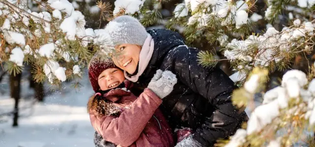 Uśmiechnięta mama i córka przytulają się w śnieżnym lesie, ciesząc się wspólnymi feriami zimowymi.
