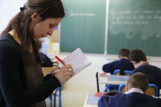 Nauczycielka notuje w zeszycie, podczas gdy uczniowie siedzą w ławkach. Temat zarobków nauczycieli jest ważny dla przyszłości edukacji.